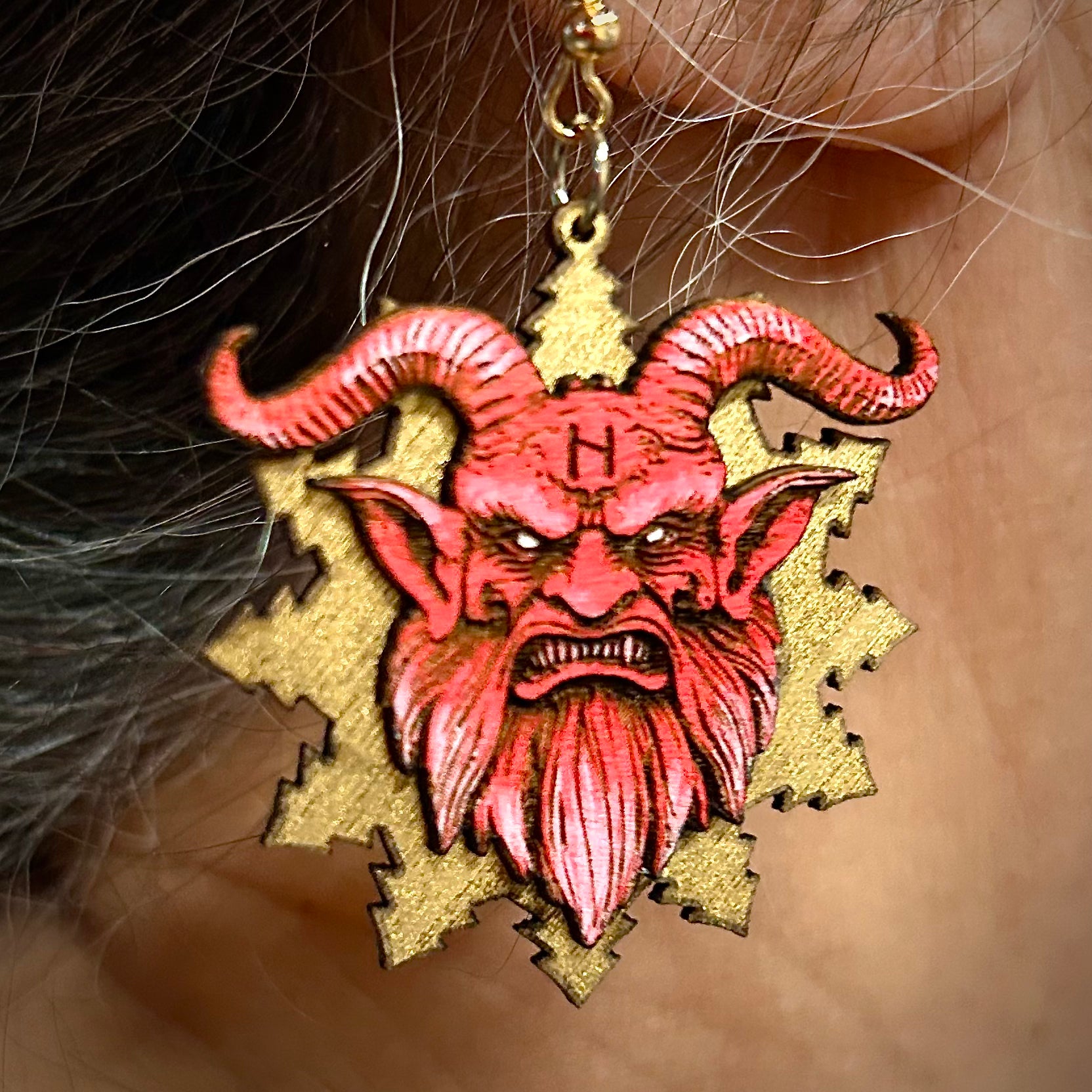 Earring with red demon face design on a gold background worn by a person.