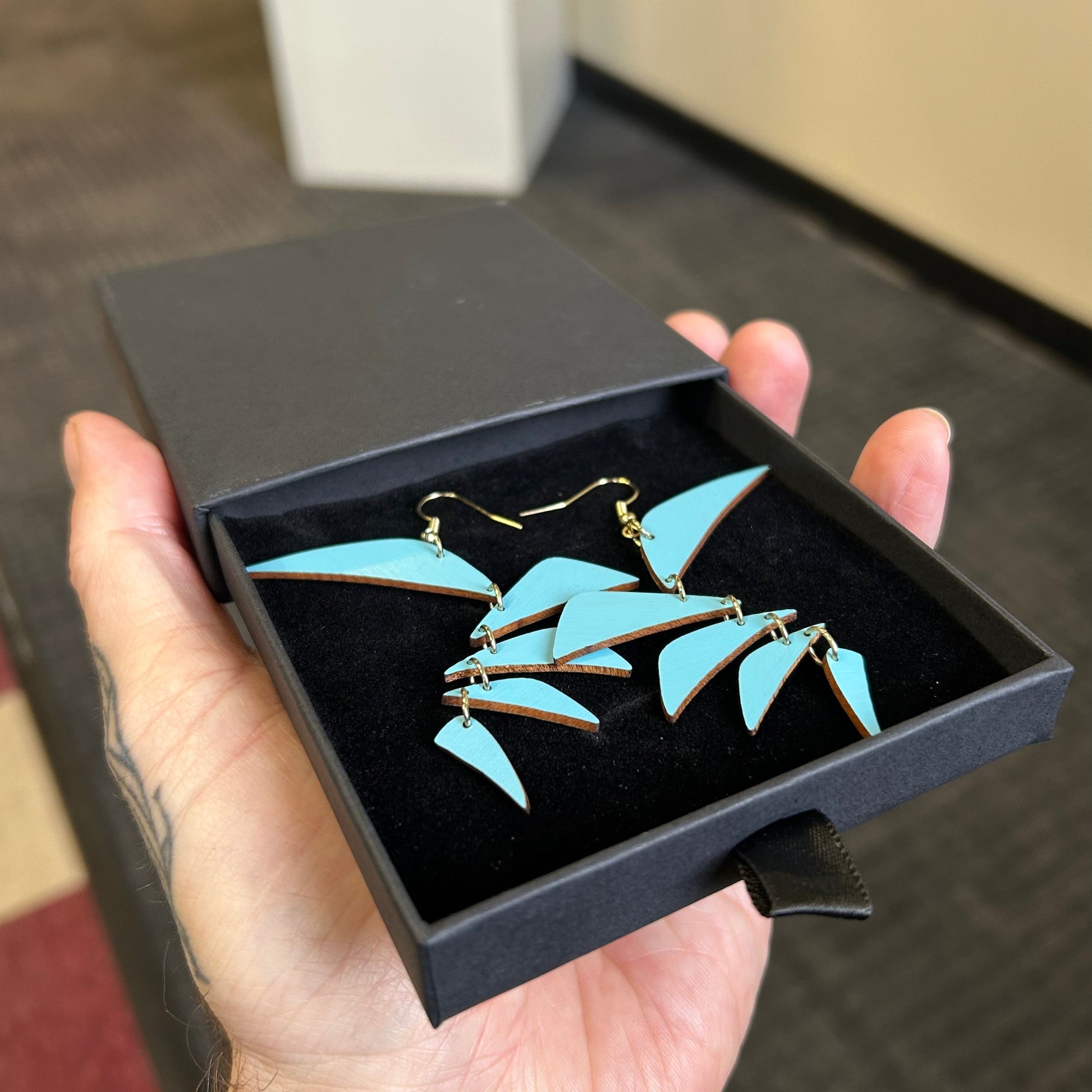 Turquoise leaf-shaped earrings in a black jewelry box held by a hand.