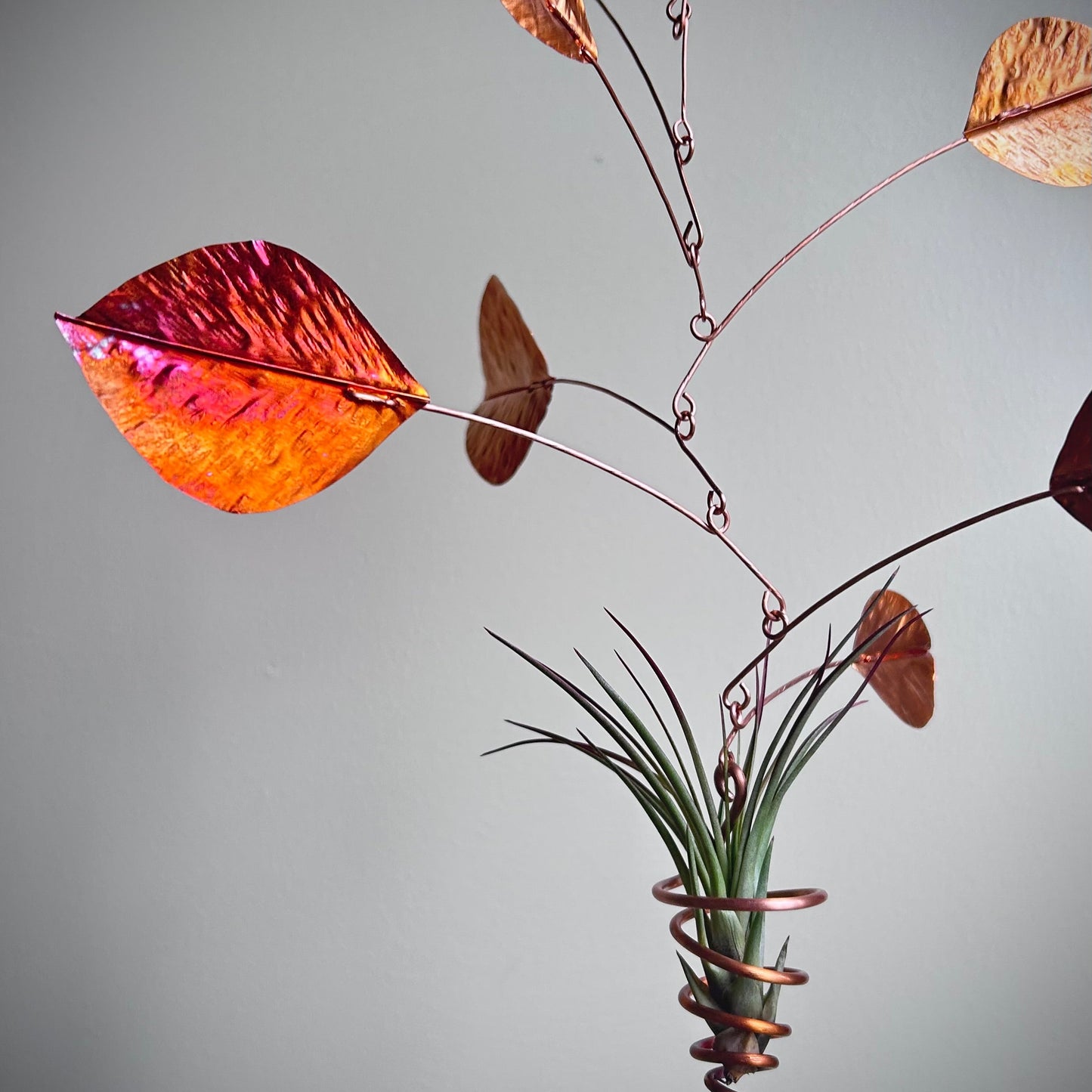 A decorative hanging wind spinner featuring copper-colored metal branches, iridescent orange and pink leaves, and a small green air plant wrapped in a copper spiral, shown against a neutral gray background.