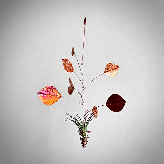 A hanging metal leaf wind spinner with copper-colored leaves, a dark burgundy leaf, and a small potted air plant at the center, suspended against a plain light gray background.